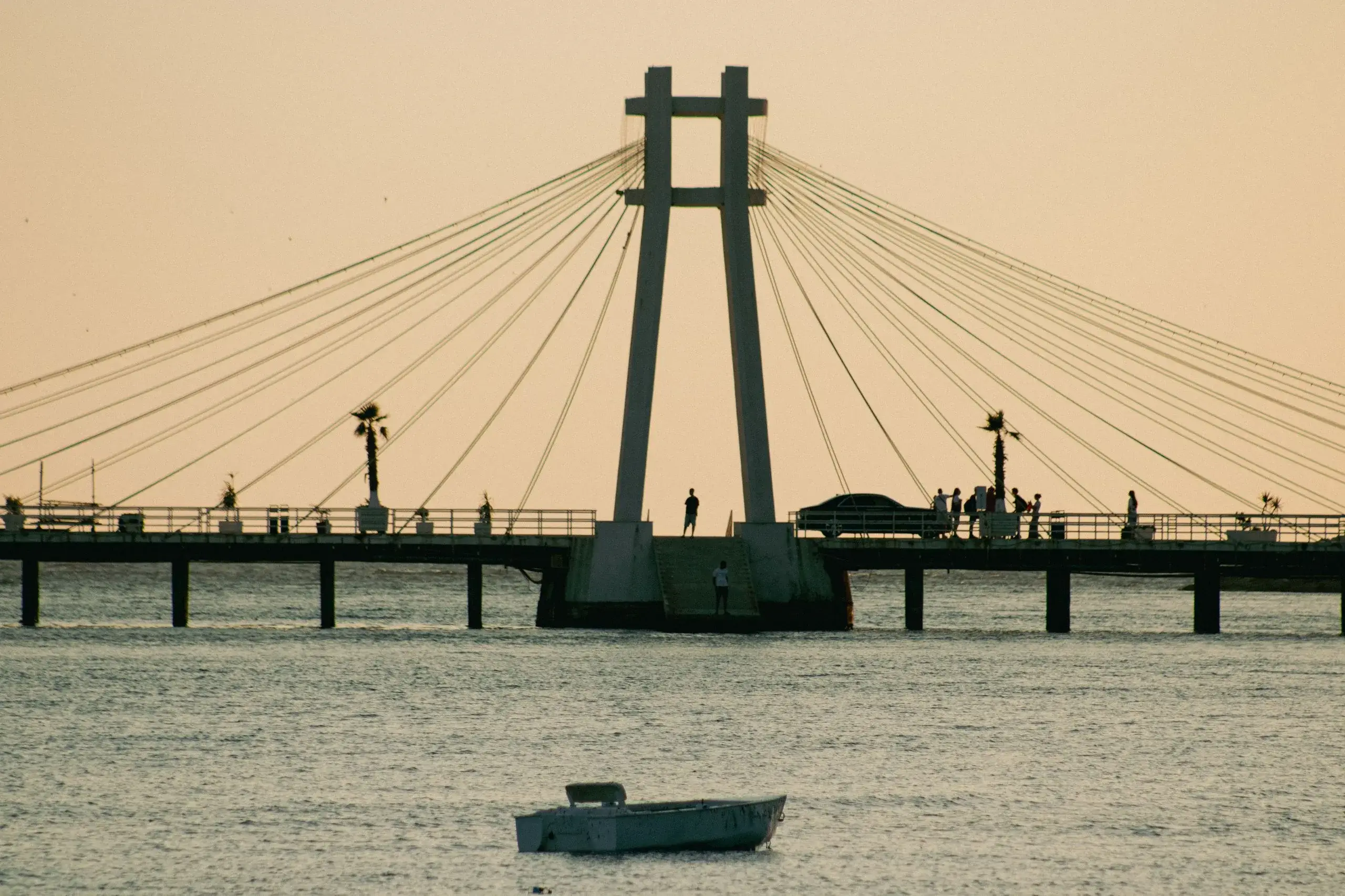 The Durrës suspension pier at sunset, with palm trees, small figures on the deck and a rowboat on the Adriatic