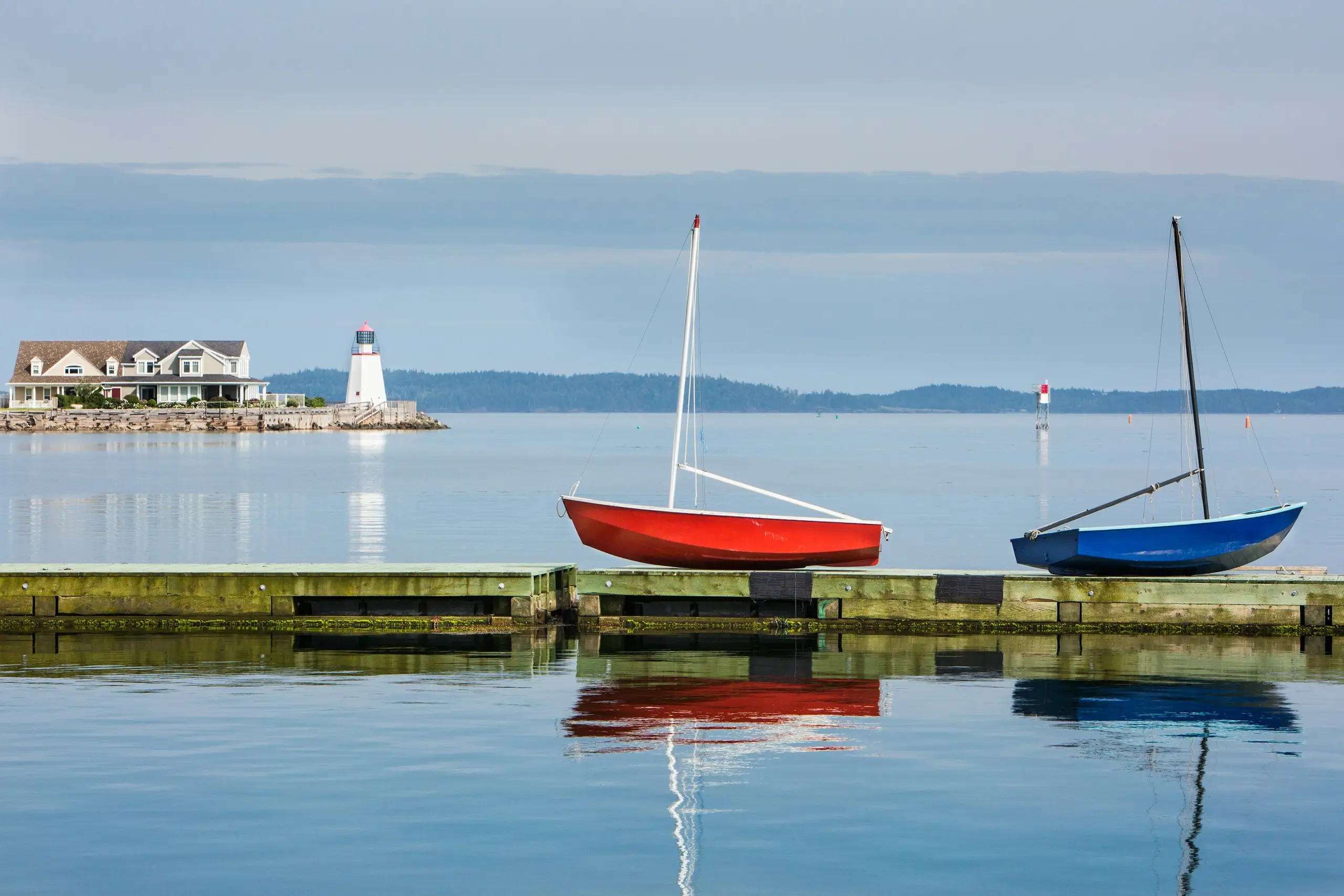 A red and a blue dinghy resting on a dock with a small lighthouse across the calm bay, Saint Andrews, New Brunswick