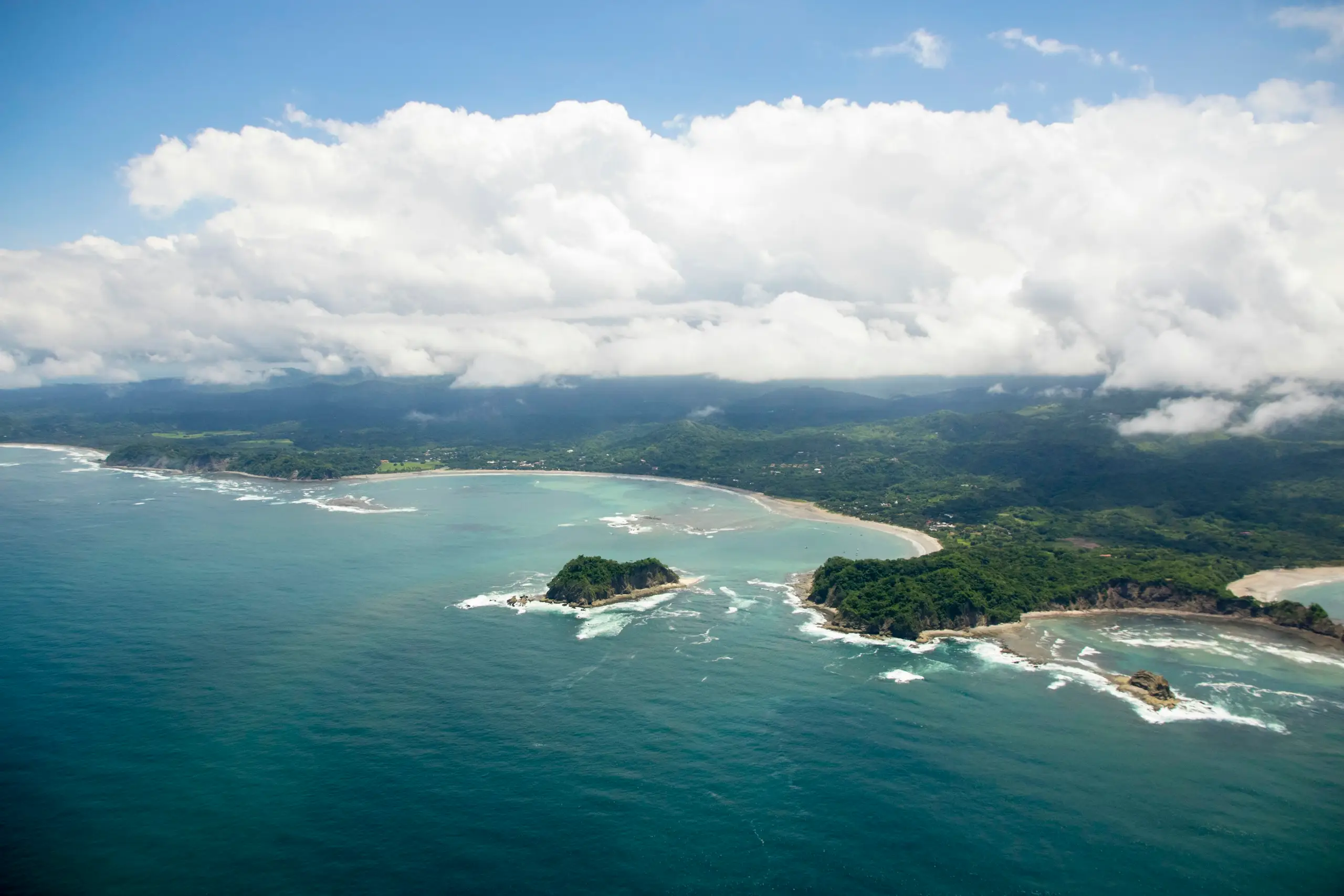 An aerial view of a curved sandy bay and offshore islet backed by green hills, Samara, Costa Rica