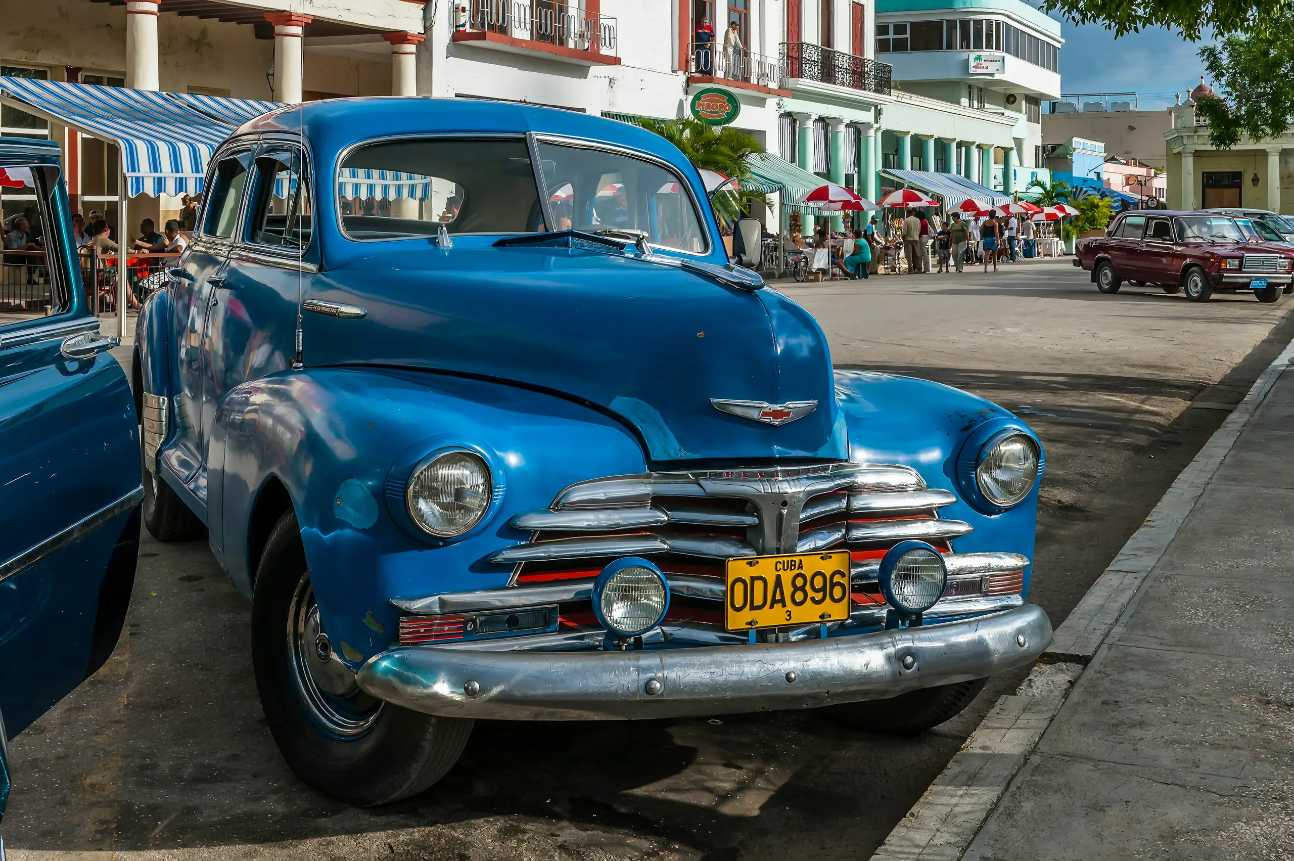 A vintage blue Chevrolet parked on a sunny street lined with cafes and parasols, Holguin, Cuba