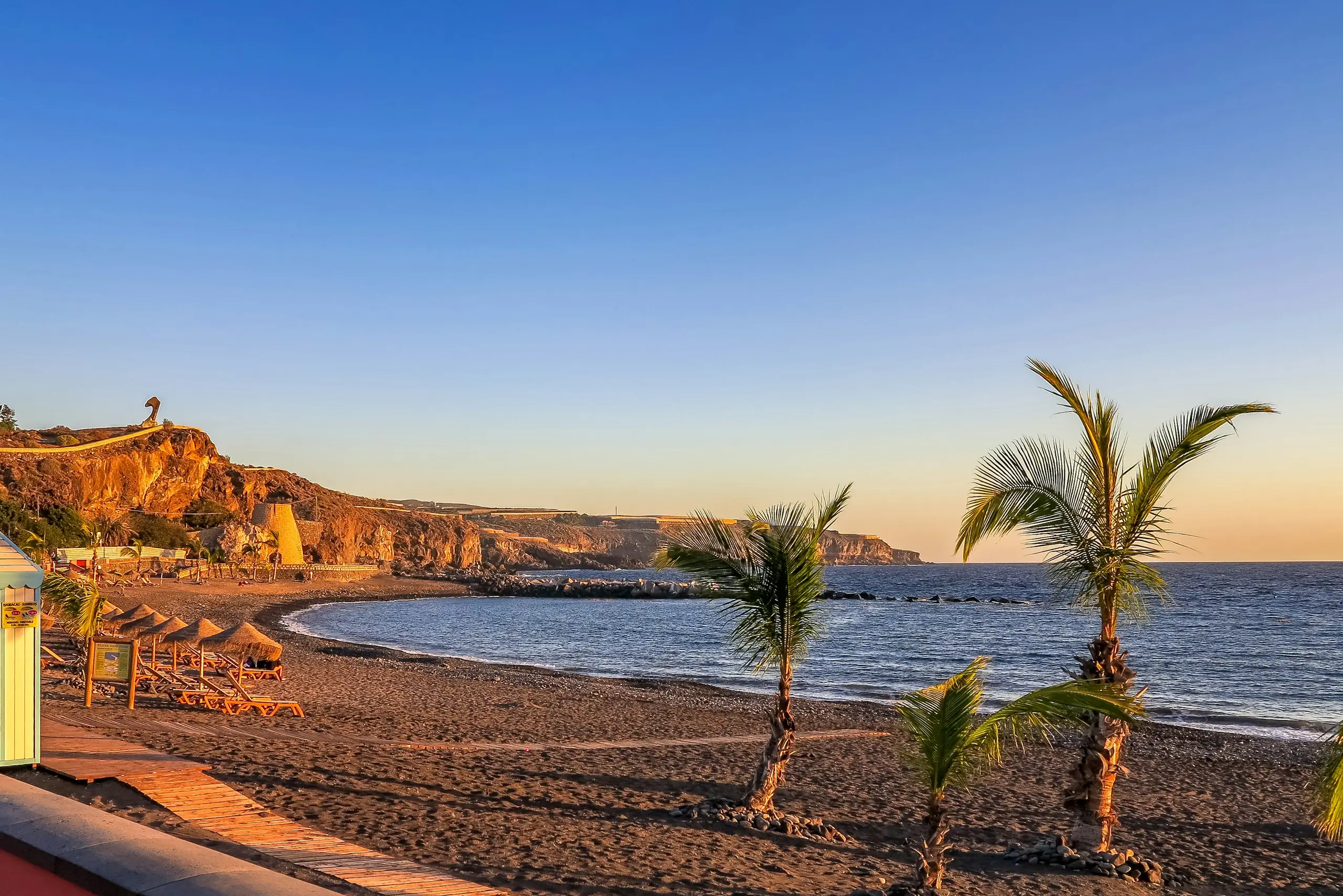 A dark-sand cove with thatched parasols and palms below golden cliffs at sunset, Tenerife, Spain