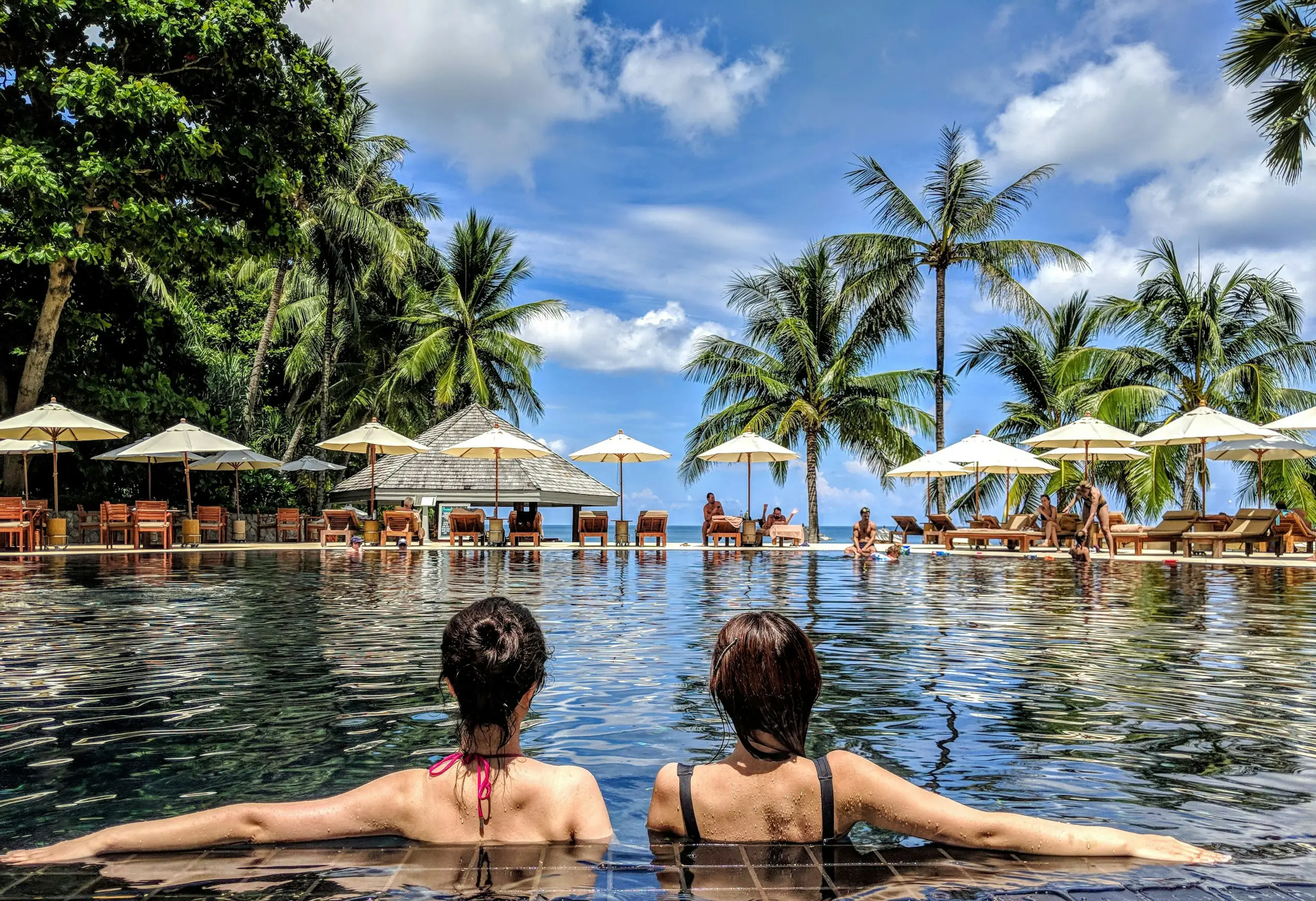 Two guests relaxing in a resort pool facing palm trees and beach umbrellas