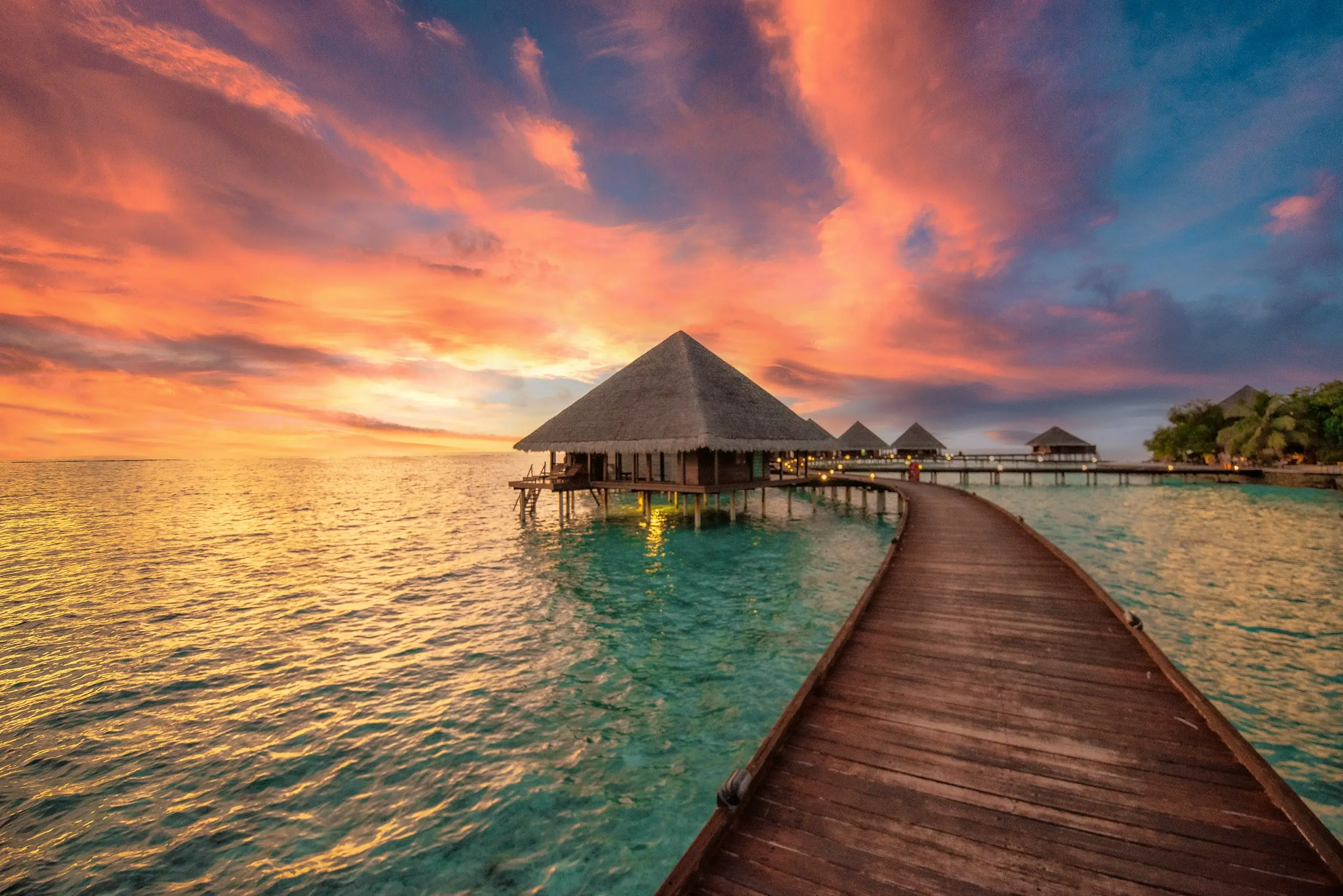 A wooden boardwalk leading to an overwater hut under a glowing orange sunset